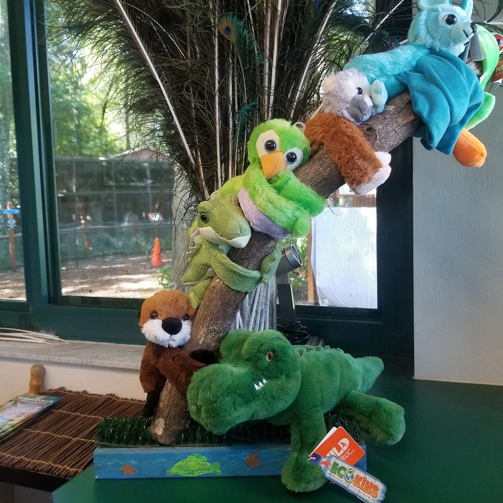 multiple stuffed animal huggers on branch with peacock feathers in background, and alligator stuffed animal