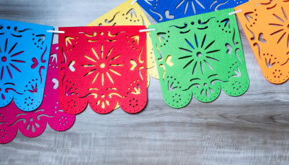 Colorful papel picado banners with floral cutout designs hanging on a string over a gray wooden background.